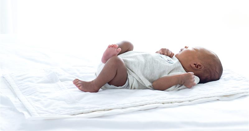 Dark-skinned baby laying on white blanket wearing a grey onesie. Dark-skinned baby laying on white blanket wearing a grey onesie.